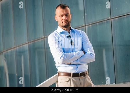 Ein Mann in blauem Hemd und khakifarbener Hose steht draußen mit überkreuzten Armen vor einem modernen Glasgebäude. Stockfoto