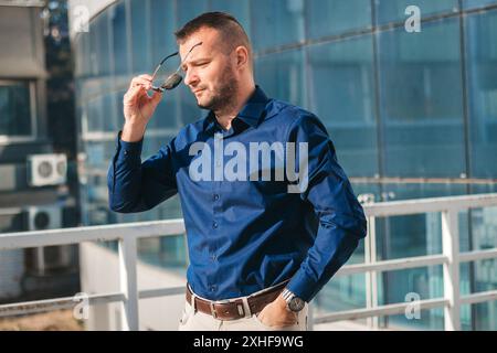 Ein Mann in einem blauen Hemd passt seine Sonnenbrille an, während er auf einem modernen Balkon steht. Stockfoto