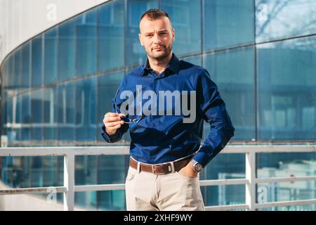 Ein Mann in blauem Hemd und khakifarbenen Hosen steht auf einem Balkon mit einem modernen Glasgebäude im Hintergrund. Er hält die Brille in seiner rechten Hand und Stockfoto