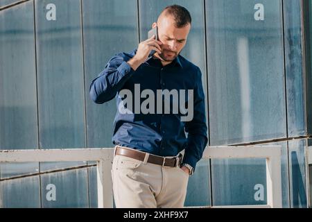 Ein Mann mit kurzen braunen Haaren und Bart, der ein blaues Hemd und eine khakifarbene Hose trägt, benutzt ein Smartphone, während er vor einem Glasgebäude steht. Stockfoto