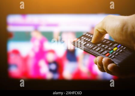Eine Hand drückt eine Taste auf einer schwarzen Fernbedienung, wobei ein unscharfer Fernsehbildschirm im Hintergrund angezeigt wird. Stockfoto