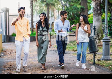 Fröhliche, abwechslungsreiche Gruppe junger Erwachsener, die gemeinsam in einem üppigen Stadtpark spazieren gehen. Sie führen animierte Gespräche, Lachen und Gesten. Die im Stockfoto