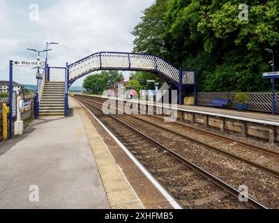 Arnside Cumbria UK. Ein malerischer ländlicher Bahnhof mit einer blauen Fußgängerbrücke, umgeben von üppigem Grün und leeren Bahngleisen an einem bewölkten Tag. Stockfoto