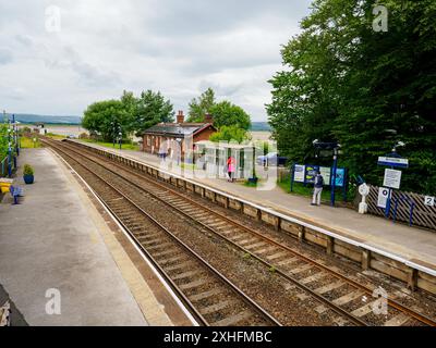 Arnside Cumbria UK. Ruhiger ländlicher Bahnhof mit Passagieren, umgeben von üppigem Grün und offenem Himmel. Stockfoto