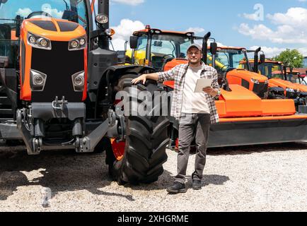 Kaukasischer Erwachsener mit digitalem Tablet in der Hand steht neben einem Bau- oder landwirtschaftlichen Traktor bei einem Maschinenhändler. AG-Gerätehändler oder -Zulieferer Stockfoto