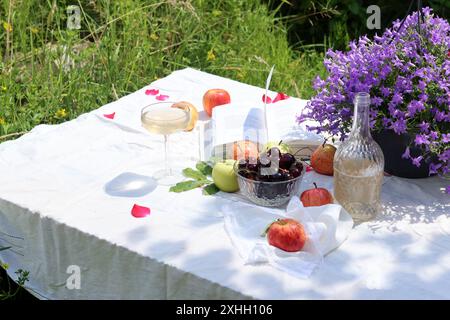 Sommer-Picknick im Garten auf einer weißen Tischdecke mit frischem Obst, Blumen und Champagner. Warmer Tag in einem Garten. Romantischer Esstisch. Stockfoto