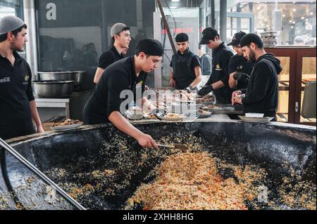 Männliche Köche legen fertige traditionelle usbekische Pilaw aus einem großen Kessel auf Teller im zentralasiatischen Pilaw Center Besh Qozon. Taschkent, Usbekistan - 16. April 2024 Stockfoto