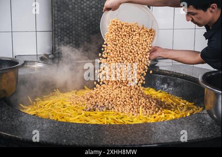 Männlicher Koch gießt Kichererbsen in Kessel mit gelben Karotten zum Kochen von usbekischen Pilaw im zentralasiatischen Pilaw-Zentrum Besh Qozon. Taschkent, Usbekistan - 16. April 2024 Stockfoto