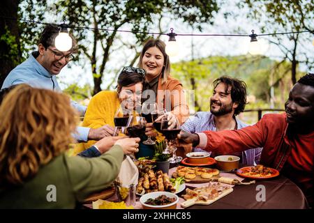 Fröhliche, vielfältige Gruppe von Freunden, die bei einer Dinner-Party im Freien einen Wein trinken. Multikulturelles Treffen mit Tapas und Lichterketten im Garten. Ce Stockfoto