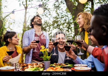 Ekstatische Gruppe multikultureller Freunde, die sich über Wein lustig lachen. Frühlingsparty im Freien mit Lichterketten, die echte Momente der Freude und festhalten Stockfoto