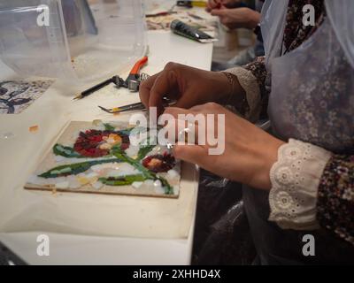 Mosaic Workshop: Ein Nahaufnahme der horizontalen Tischplatte fängt zwei Paar Frauenhände ein, die während der Mos Blumenmosaikmuster auf dem weißen Tisch basteln Stockfoto