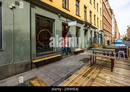 Die Bar Udda hat ihre grün gestrichene Fassade an der Kocksgatan in Södermalm, Stockholm – dem coolsten Viertel der Stadt, mit Vintage-Läden, Galerien, Bars, Restaurants und Bio-Cafés, die in ehemaligen Arbeiterviertel-Hausblocks untergebracht sind. Udda vereint eine Bar, eine kleine Brauerei und eine Waschküche unter einem Dach. Die Journalistin Angela Berg steht im rot gestrichenen Eingang, mit Holzbank und Tisch auf der Holzterrasse davor. Kocksgatan, Södermalm, Stockholm, Stockholms län, Schweden. Stockfoto