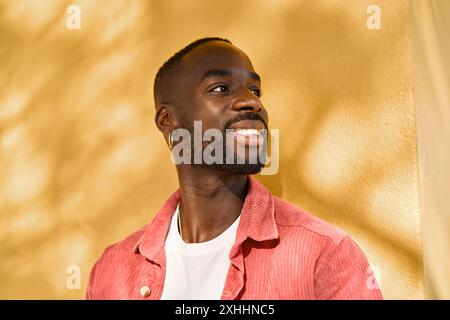 Der lächelnde coole Gen z Schwarzer Hipster-Typ sieht weg und steht an der gelben Wand. Stockfoto
