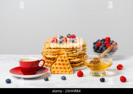 Belgische Waffeln, Beeren, Honig und eine rote Tasse Kaffee: Köstliches Sommerfrühstück auf Marmortisch. Herzförmige Waffel und eine Mischung aus Himbeeren Stockfoto