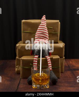 Ein Weihnachtszwerg sitzt auf einem Geschenk und trinkt Whiskey durch einen Strohhalm. Schwarzer Hintergrund, Holzboden, Präsentationen in Recyclingpapier verpackt Stockfoto