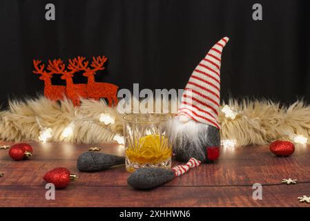 Ein Weihnachtszwerg, der auf einem Holzboden sitzt, mit einem fast leeren Glas Whiskey. Im Hintergrund drei rote Rentiere, Feenlichter, Herzen und Stern Stockfoto