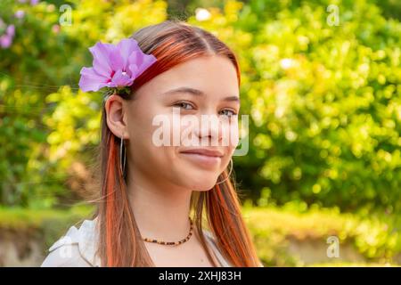 Ein Mädchen im Teenageralter mit langen rötlich-braunen Haaren lächelt in die Kamera, mit einer rosa Blume hinter ihrem Ohr. Sie trägt ein weißes Hemd und einen Halsausschnitt Stockfoto