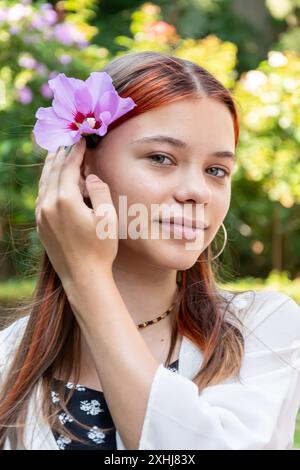 Ein 13-jähriges Mädchen steckt vor dem Hintergrund eines blühenden Gartens eine Blume ins Haar. Stockfoto