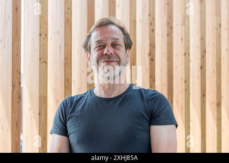 Ein Mann mit einem 50-55-jährigen Bart lächelt vor einer Holzwand in die Kamera. Stockfoto