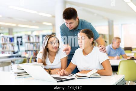 Junger Mann in lässiger Kleidung, der jungen Frauen, die auf dem Bildschirm des Computers in der Bibliothek pochieren, Informationen erklärt Stockfoto