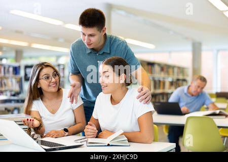 Junger Mann in lässiger Kleidung, der jungen Frauen, die auf dem Bildschirm des Computers in der Bibliothek pochieren, Informationen erklärt Stockfoto