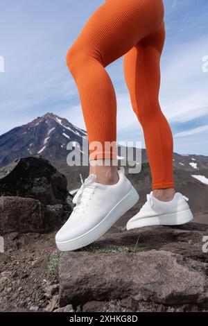 Sportliche weibliche Beine mit orangefarbenen Leggings und Sneakers. Nahaufnahme des unteren Teils des Körpers der Frau, während sie draußen in Bergen mit Sport treibt Stockfoto
