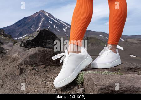 Nahaufnahme von Frauenbeinen mit weißen Turnschuhen und orangefarbenen Sportleggings. Frau steht während des Fitnesstrainings in den Bergen mit wunderschöner Aussicht Stockfoto