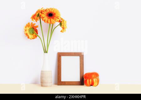 Leerer Bilderrahmen, orange Gerbera-Blüten in Vase und Kürbis auf dem Tisch. Modell Stockfoto