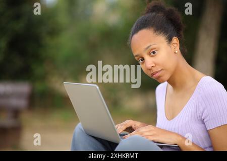 Eine ernste schwarze Frau, die einen Laptop benutzt, um in einem Park in die Kamera zu schauen Stockfoto