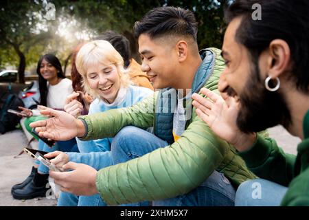 Eine multiethnische Gruppe junger Erwachsener genießt die gemeinsame Zeit im Freien. Freunde lachen und teilen Inhalte auf dem Smartphone. Verschiedene Millennials verbinden sich über Stockfoto