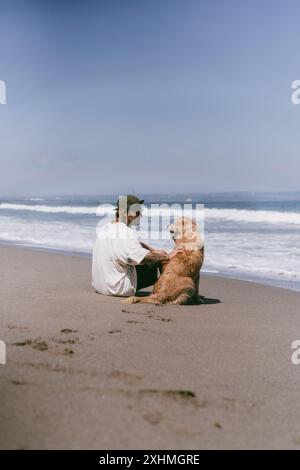 Der Mann sitzt am Meer am Strand mit goldenem Retriever-Hund. Stockfoto