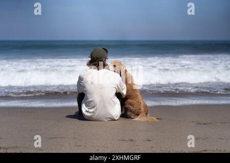 Der Mann sitzt am Meer am Strand mit goldenem Retriever-Hund. Stockfoto