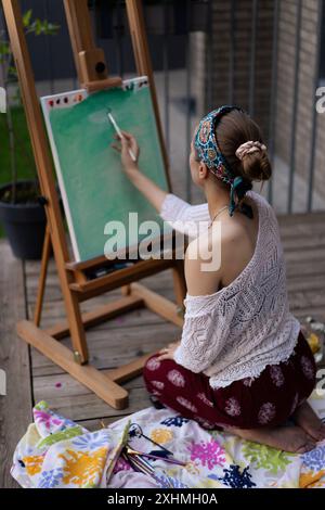 Frau Künstlerin malt auf Leinwand, Ölfarben. Staffelei auf Terrasse, Sommer. Stockfoto