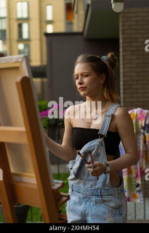 Frau Künstlerin malt auf Leinwand, Ölfarben. Staffelei auf Terrasse, Sommer. Stockfoto