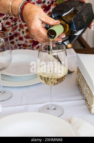 Eine Nahaufnahme einer Hand, die an einem schön gedeckten Esstisch Weißwein in ein Glas gießt. Die Szene fängt die Eleganz und Erwartung einer Flosse ein Stockfoto