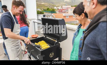 Fröhliche Freunde kochen Mais beim Barbecue-Abendessen auf dem Dach - Mehrgenerationenmenschen, die im Sommer Spaß beim Essen und Lachen haben - Main Foc Stockfoto