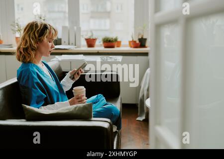 Profilaufnahme einer blonden, kaukasischen Krankenschwester, die einen Thermobecher hält und ihre sozialen Netzwerke überprüft, während sie während ihrer Pause in Lotusposition auf weichem Sofa sitzt Stockfoto