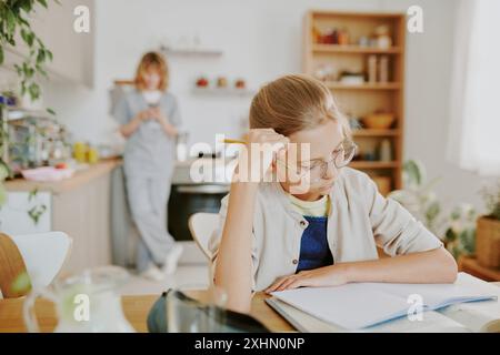 Mittlere Nahaufnahme eines goldhaarigen kaukasischen Mädchens, das Buch liest, während ihre Ärztin Mom das Telefon in unscharfem Hintergrund überprüft Stockfoto