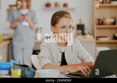 Mittlere Nahaufnahme eines hübschen kaukasischen Mädchens, das Hausaufgaben über einen schwarzen Laptop macht, während ihre Mutter die Küchenzeile lehnt und das Telefon im unscharfen Hintergrund überprüft Stockfoto