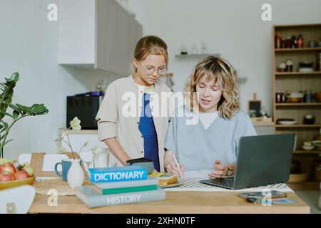 Die junge Ärztin sitzt am Tisch und erklärt Hausaufgaben Fehler, während ihre Tochter neben ihr steht und aufmerksam zuhört Stockfoto