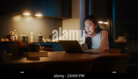 Schöne authentische asiatische Frau, die an einem Tisch in einer gemütlichen Küche sitzt und nachts einen Laptop benutzt. Frau lächelt und chattet im Internet und in sozialen Medien, forscht Stockfoto