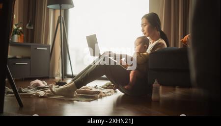 Arbeiten von zu Hause aus: Porträt der jungen asiatischen Mutter, die ihr süßes Baby hält und tagsüber an einem Laptop arbeitet. Erfolgreiche weibliche Führungskraft, die Leben und Arbeit in Einklang bringt, während sie ihre Arbeit aus der Ferne erledigt Stockfoto