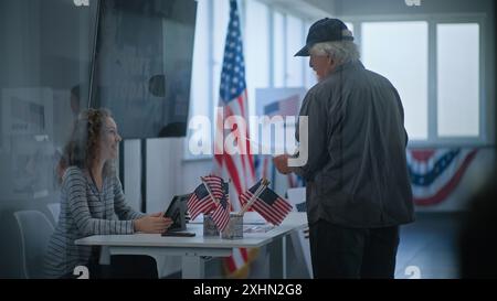 Kaukasischer reifer US-Bürger steht in der Nähe der Registrierungstafel am Wahlschalter. Die weibliche Wahlverantwortliche spricht mit dem Mann, benutzt einen Tablet-Computer und gibt Stimmzettel für die Wahl. Wahltag in den USA. Stockfoto
