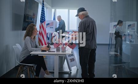 Kaukasischer reifer US-Bürger steht in der Nähe der Registrierungstafel am Wahlschalter. Die weibliche Wahlverantwortliche spricht mit dem Mann, benutzt einen Tablet-Computer und gibt Stimmzettel für die Wahl. Wahltag in den USA. Stockfoto