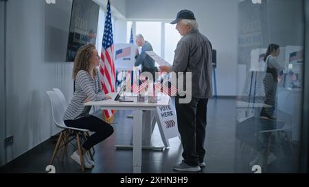 Kaukasischer reifer US-Bürger steht in der Nähe der Registrierungstafel am Wahlschalter. Die weibliche Wahlverantwortliche spricht mit dem Mann, benutzt einen Tablet-Computer und gibt Stimmzettel für die Wahl. Wahltag in den USA. Stockfoto