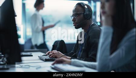 Porträt eines Smart African Young Specialist, der in einer technischen Startup-Helpdesk-Abteilung arbeitet und Computer in einem vielfältigen Büro verwendet. Schwarzer Mann, der Brille und Kopfhörer mit Mikrofon trägt Stockfoto