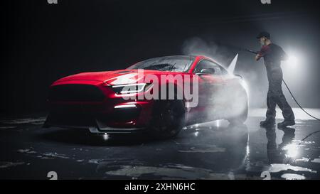 Erwachsenenautos in Uniform waschen einen roten Sportwagen mit einem Hochdruckreiniger. Reinigungstechniker, der an einem stilvollen amerikanischen Auto in einem dunklen Raum arbeitet. Kommerzielles Studio-Foto für Werbung Stockfoto