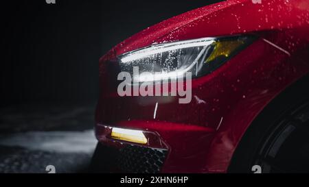 Modernes Red Performance Fahrzeug mit LED-Scheinwerfern wird in einer Autohändler-Waschanlage gereinigt. Nahaufnahme eines Werbefotos eines schnellen Autos, das in einem Low Key Cinematic Studio gewaschen wird Stockfoto