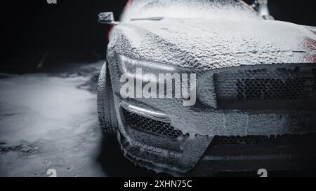 Moderne Red Performance Fahrzeugscheinwerfer, die mit Shampoo bedeckt sind, werden in einer Autowaschanlage eines Autohauses gereinigt. Nahaufnahme eines Werbefotos eines schnellen Autos, das in einem Low Key Cinematic Studio gewaschen wird Stockfoto