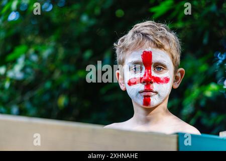 Ein hellhaariger weißer Junge mit blauem Auge, dessen Gesicht mit dem rot-weißen Kreuz von St. George bemalt ist, der die englische Fußballmannschaft unterstützt, stellt Augenkontakt her Stockfoto